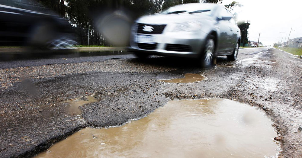 A car approaches a large pothole filled with muddy water.