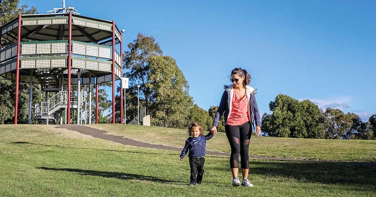 A woman and young child walk hand in hand across a grassy park on a sunny day, with a multi-level lookout tower and trees in the background under a clear blue sky.
