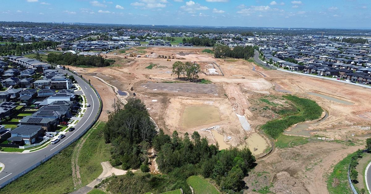 Aerial view of a large residential development site showing extensive earthworks, drainage basins and construction activity, surrounded by established housing estates and road networks under a clear blue sky.