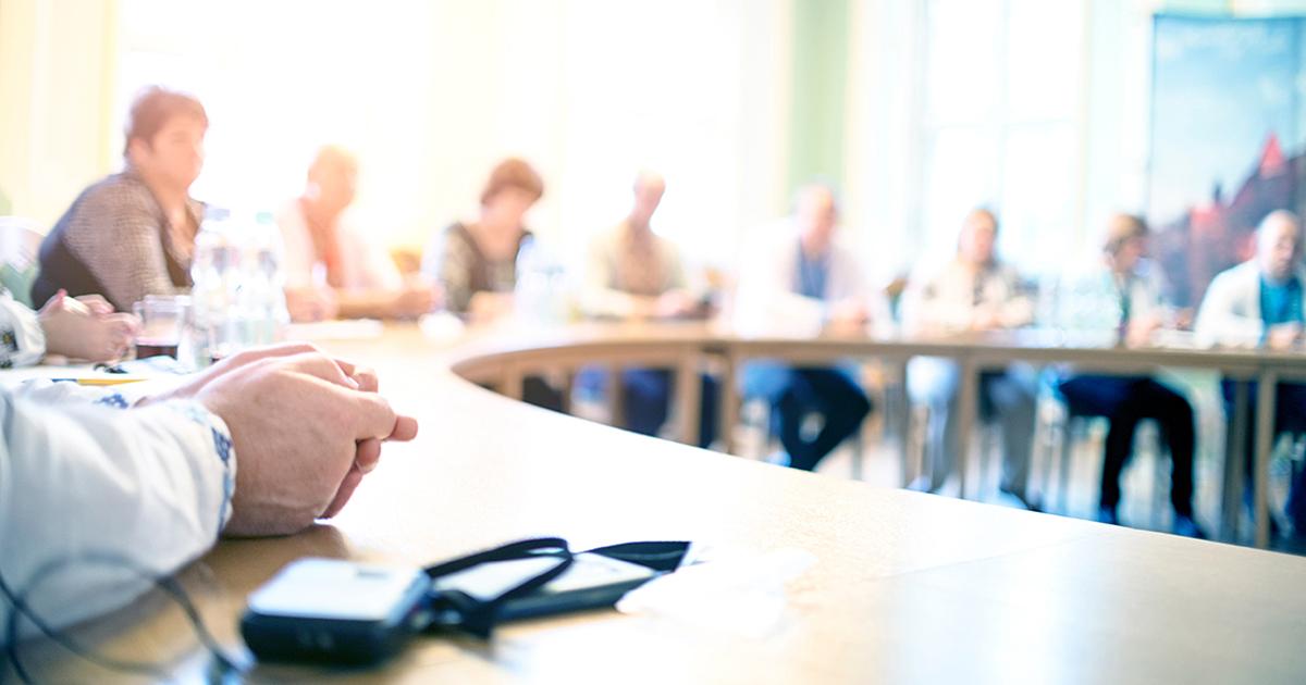 People seated around a meeting table during a council or committee discussion, with the foreground in focus and participants blurred in the background.