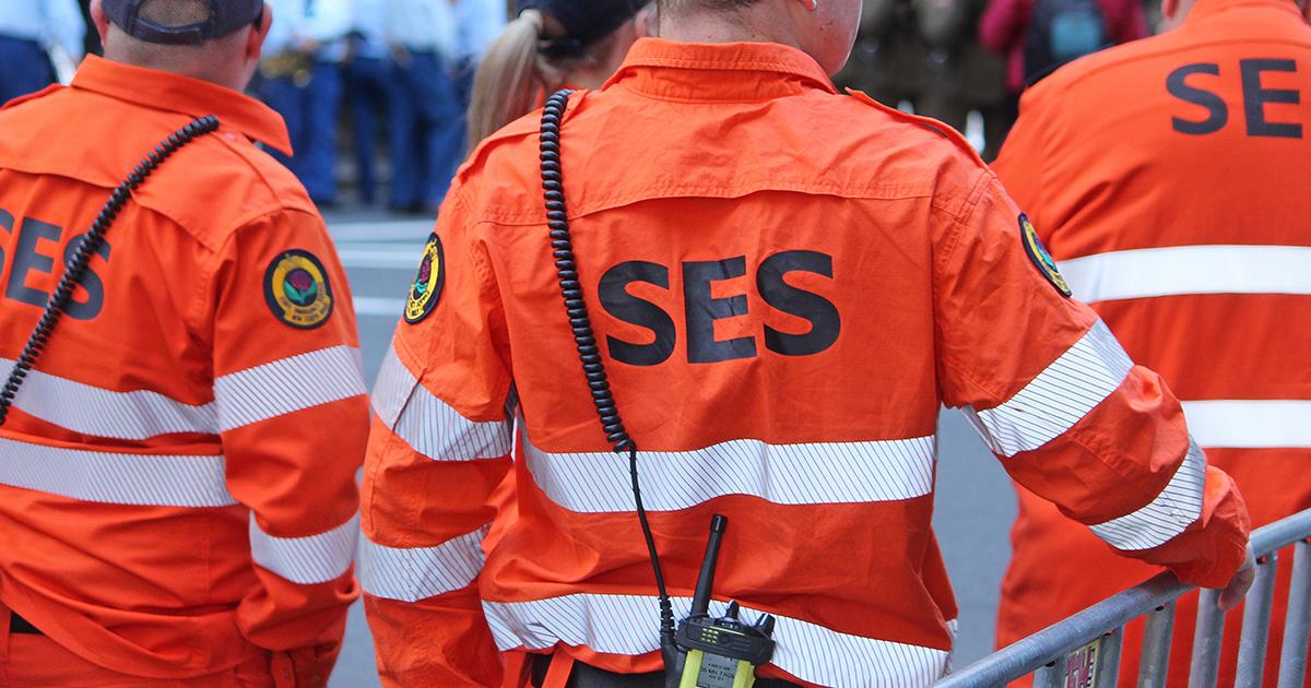 NSW SES volunteers in high-visibility orange uniforms stand ready at a community event, with radios and safety equipment visible.