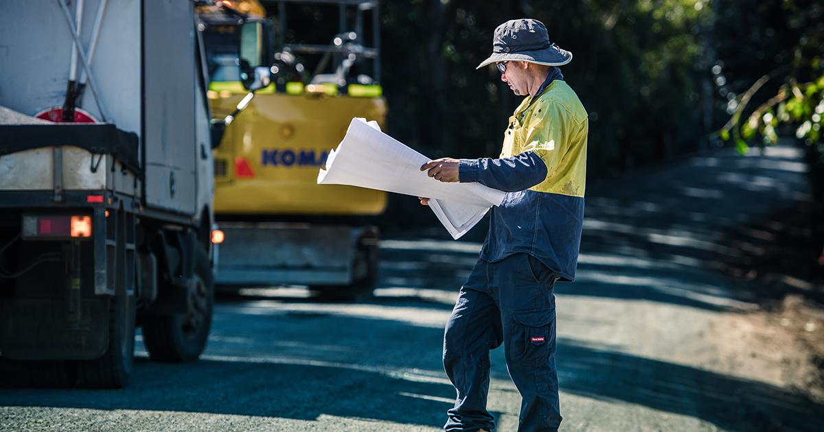 A council road worker in high-visibility clothing reviews construction plans beside work vehicles at a roadworks site.
