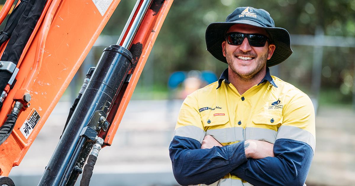 Council outdoor worker in a yellow and navy high-visibility uniform standing with arms crossed beside an orange excavator, smiling at the camera.