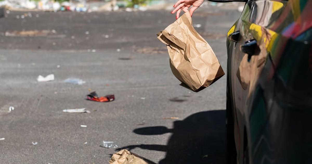 Hand dropping a paper bag from a car onto a littered roadside, highlighting illegal dumping and roadside littering.