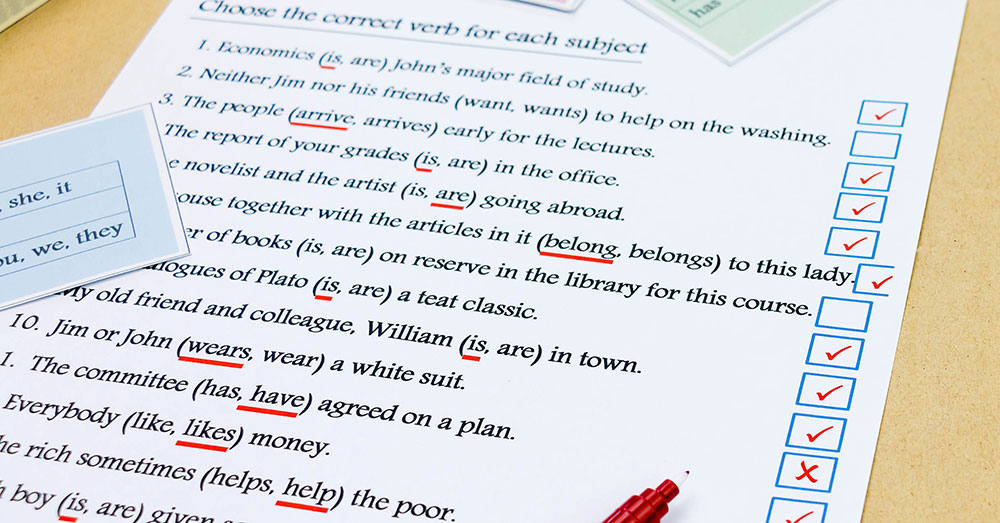 A red pen rests on a printed document that has tick boxes for correct answers to grammar questions.