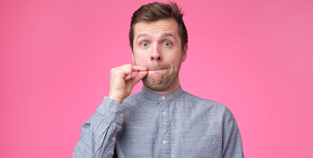 A man displays the zipping lips sign in front of a striking pink background.