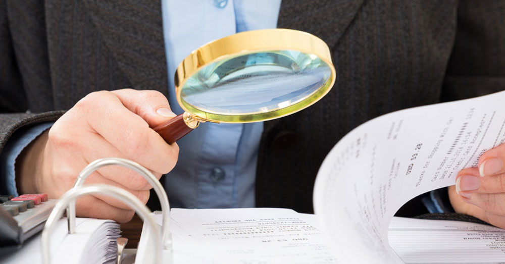 Man holding magnifying glass over document in a ring binder.