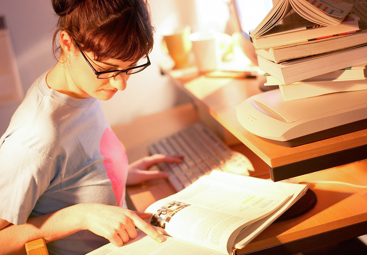 A woman at her computer with an open book and her finger placed on a section of that book.