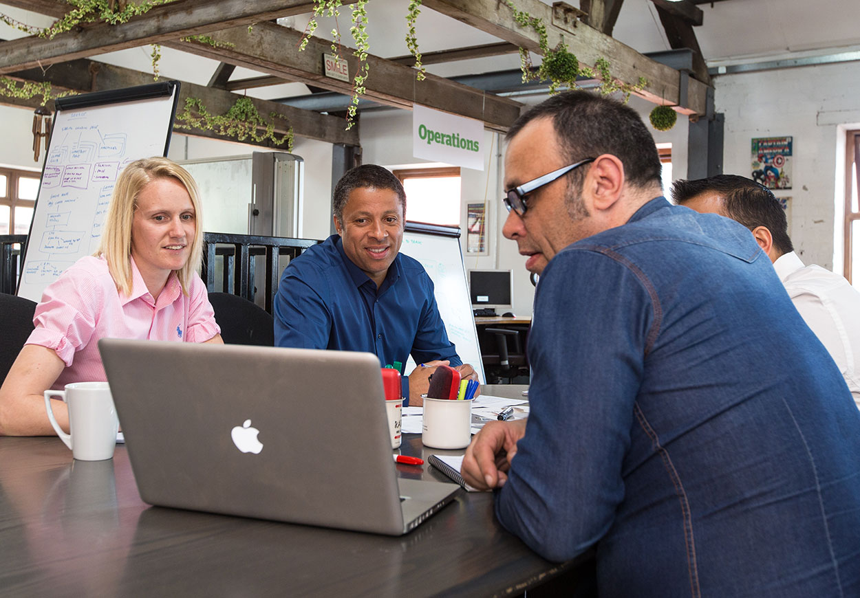 A group of four people in discussion while looking at an open Apple laptop.