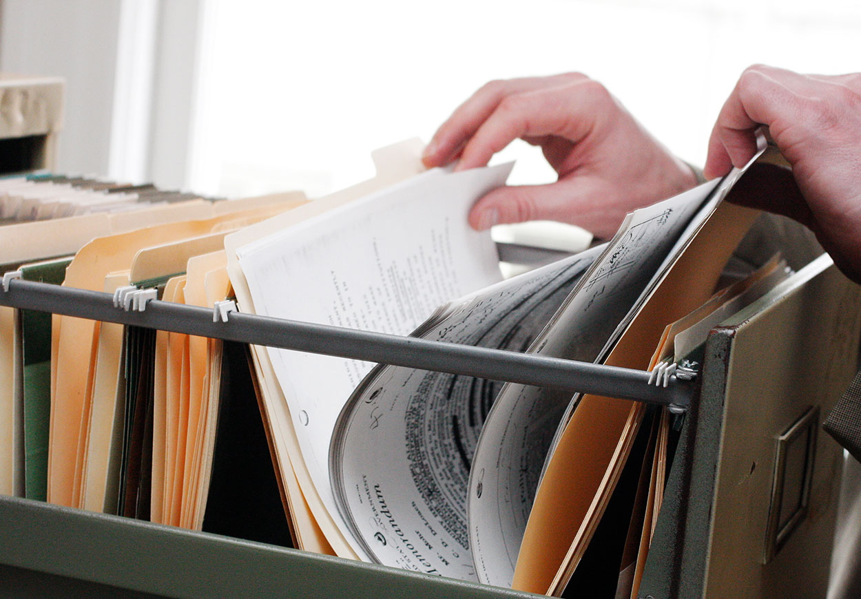 Hands thumbing through a file in an open filing cabinet drawer.