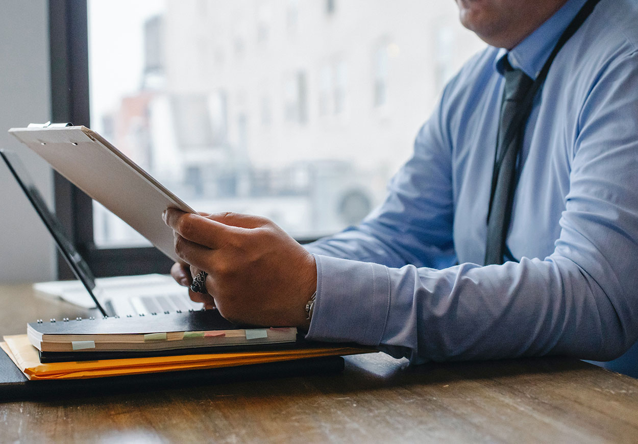 A man sitting at a desk is reading a document on a clipboard.