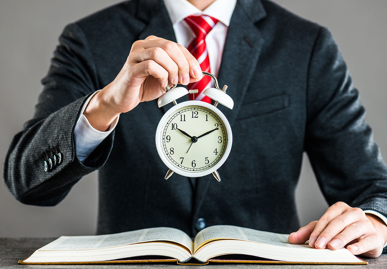 A man in a business suit and red tie holds an old-style alarm clock above an open book.