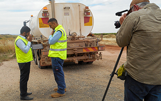 Two men in front of water truck being filmed while they are in discussion.