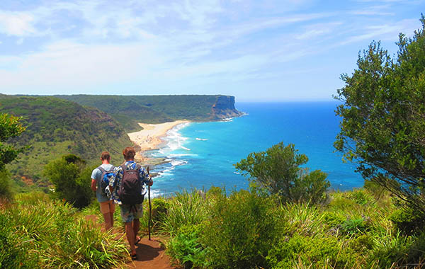 wo young men trekking through the Royal National Park, near Sydney Australia with sticks and backpacks on with a lovely view of Gerai Beach ahead on top of a hill track.